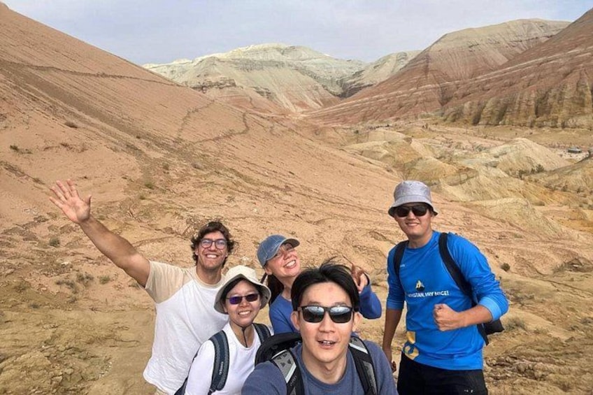Eine Gruppe Wanderer durchquert eine Wüstenlandschaft mit felsigem Gelände und Hügeln im Hintergrund. 2