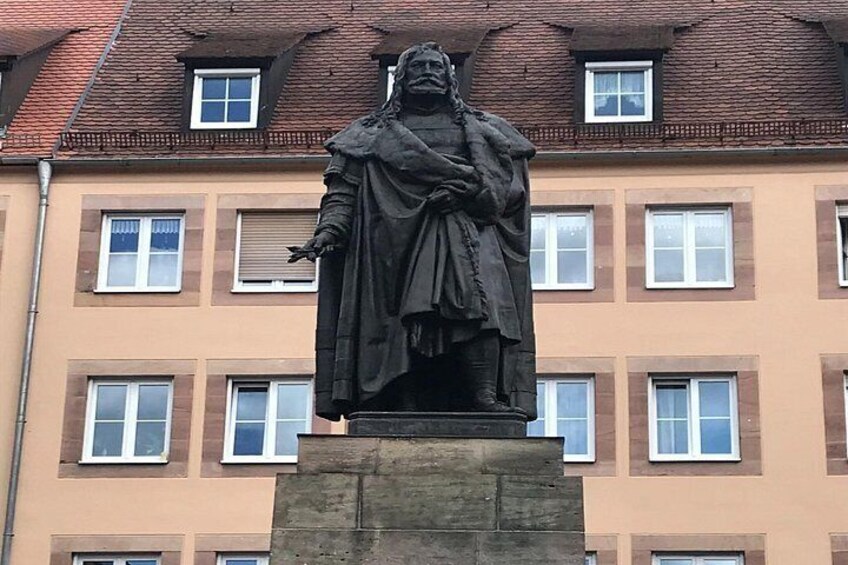 Eine Statue einer historischen Persönlichkeit in historischen Gewändern steht auf einem Sockel, im Hintergrund ist ein Gebäude mit mehreren Fenstern zu sehen. 5