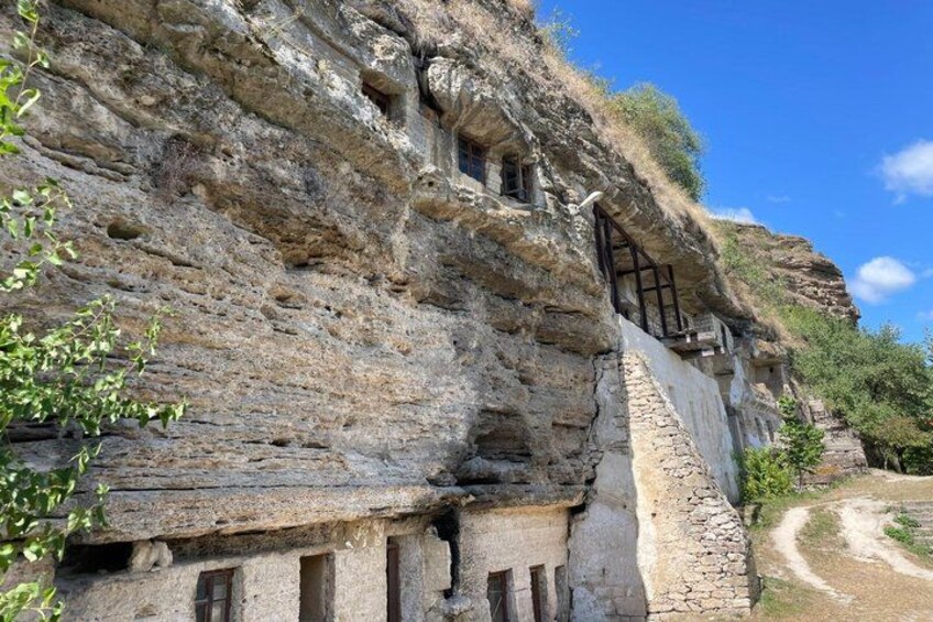 A cliffside with ancient rock-cut dwellings, a narrow path, and sparse vegetation. 1