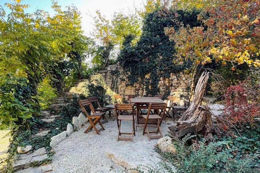 An outdoor seating area with wooden furniture, surrounded by greenery and a stone wall. 4
