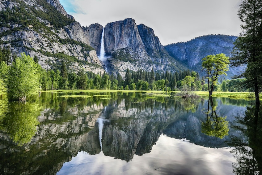 A calm lake reflecting a mountain with a waterfall, surrounded by lush green trees and a clear sky. 4