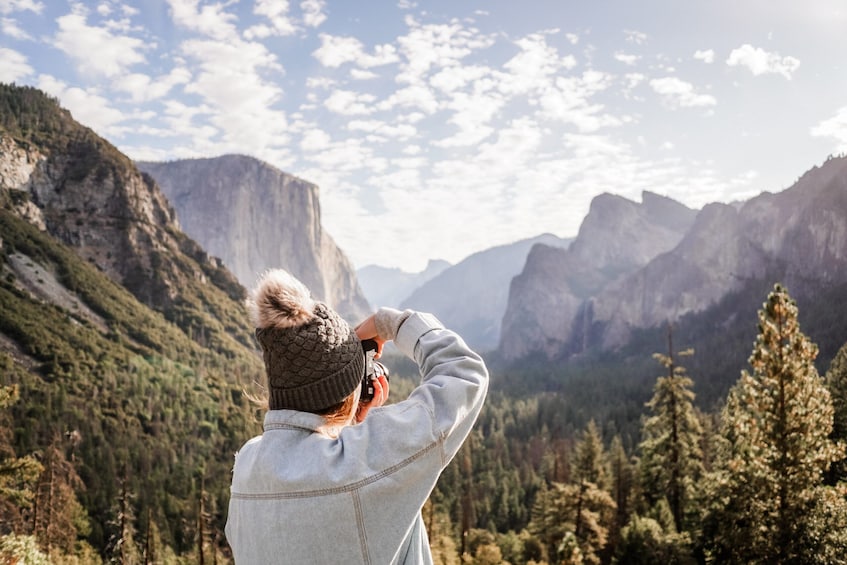 A person taking a photograph of a mountainous landscape with dense forests and rocky cliffs. 3
