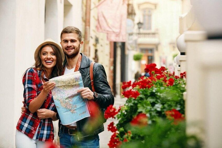 Two people holding a map, standing in a narrow street with red flowers and buildings in the background. 3