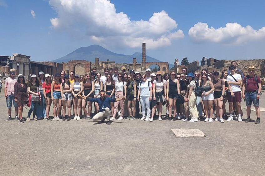 Un grupo de personas hacen fila al aire libre, con ruinas y una montaña de fondo. 1