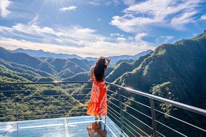 A person in an orange dress standing on a glass balcony overlooking a mountainous landscape. 2