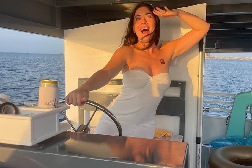 A person in a white towel on a boat, steering wheel in hand, with a can of Coca-Cola and a bottle of water on the table. 2