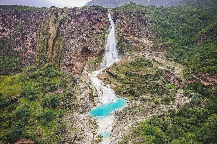 Ein Wasserfall stürzt über eine felsige Klippe in einen türkisblauen Pool, umgeben von üppiger Vegetation. 1