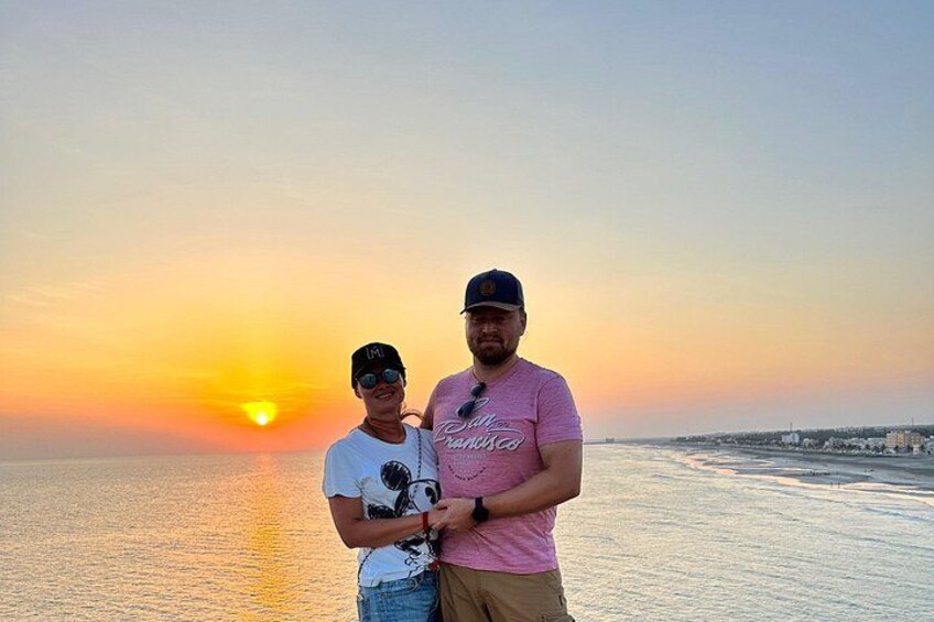 Zwei Personen stehen an einem Strand, im Hintergrund ein Sonnenuntergang. 3