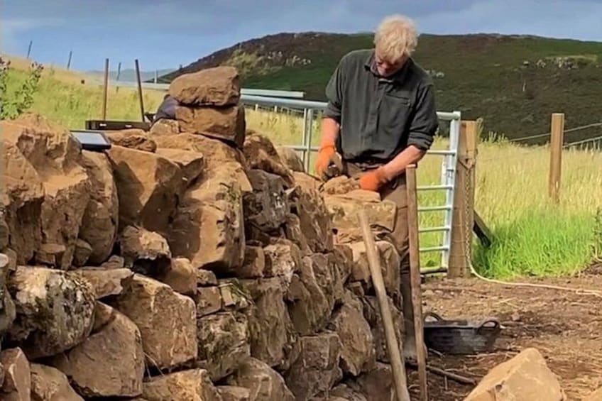 A person working with a pile of rocks, possibly for construction or landscaping, with a rural landscape and a fence in the background. 3