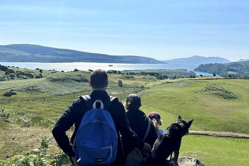 A person with a backpack and a dog on a grassy hill overlooking a body of water and distant hills. 1