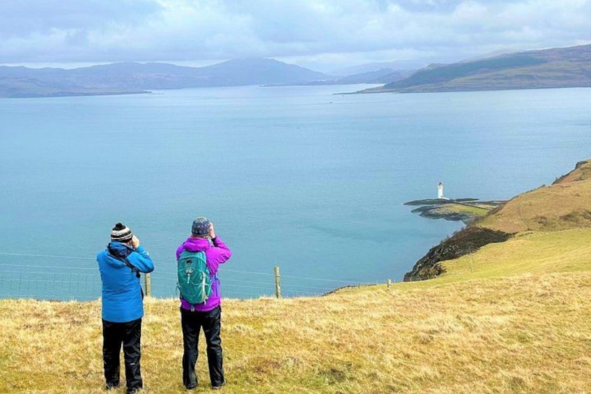 Two people standing on a grassy hill overlooking a large body of water with a lighthouse in the distance. 4