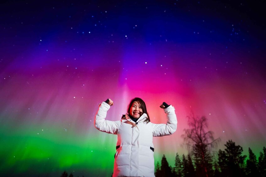 A person in a white jacket with arms raised against a colorful aurora backdrop. 1