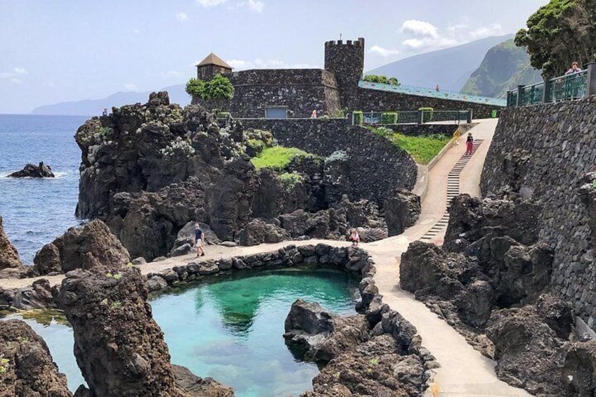 A coastal landscape with a stone pathway leading to a small pool, surrounded by rocky terrain and a historic building with a tower. 2
