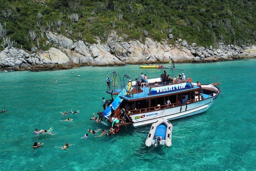 A boat with people swimming in the water, a rocky shoreline, and a clear blue sea. 2