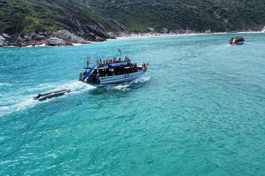 Een boot genaamd 'Volenty' op turquoise water, met een ruige kustlijn en bergen op de achtergrond. 2