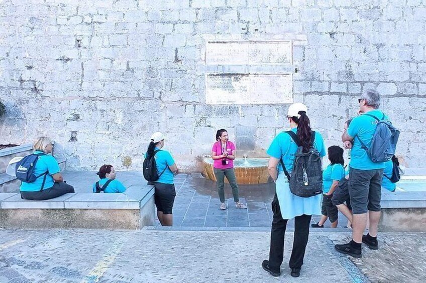 A group of people sitting and standing near a stone wall with a fountain. 1