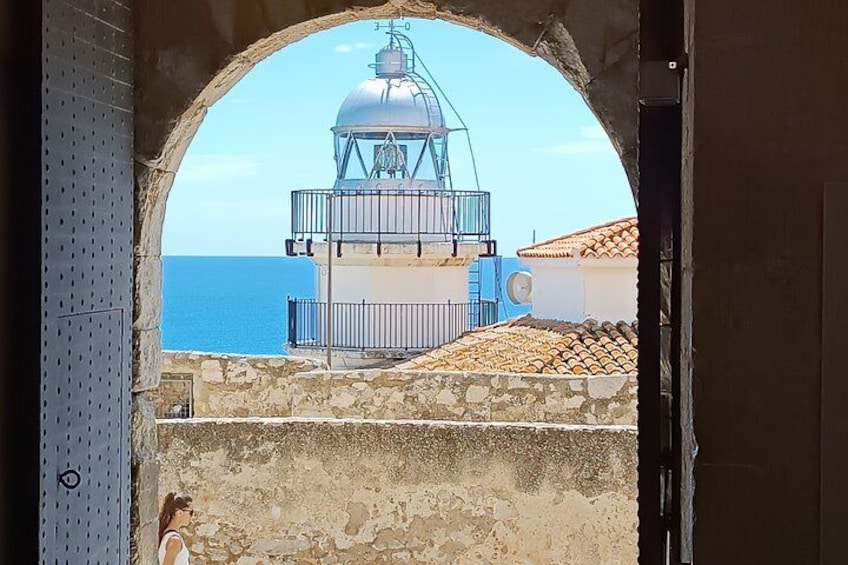 A coastal view through an archway, featuring a lighthouse, a building with a tiled roof, and the sea. 2