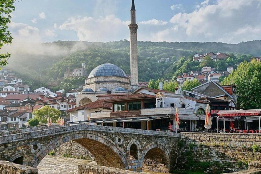 A stone bridge, a mosque with a dome, and a minaret in a town with houses and greenery in the background. 4