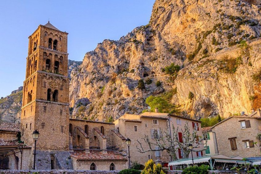 A historic stone church with a bell tower, surrounded by traditional buildings and a rocky mountain backdrop. 3