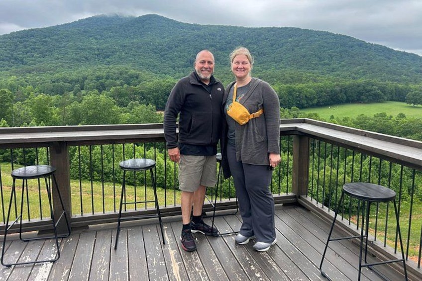 A wooden deck with metal railings, two people standing, and a view of a forested mountain range. 3
