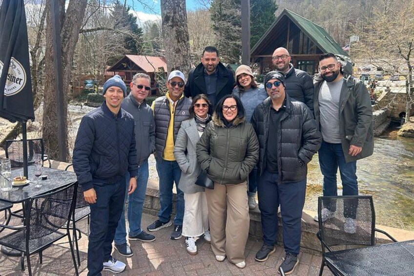A group of people standing outdoors near a table and chairs, with a wooden cabin and a river in the background. 2