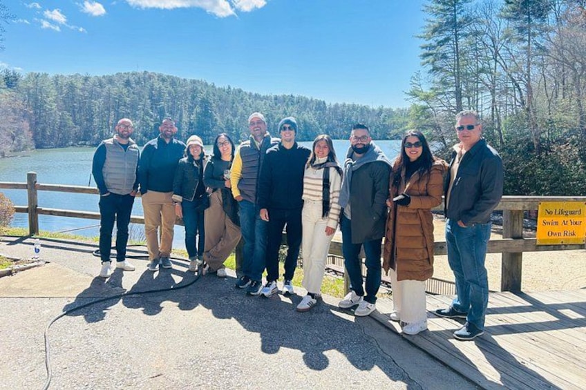 A group of people standing on a wooden boardwalk by a lake with a sign warning of no lifeguard and swim at your own risk. 2