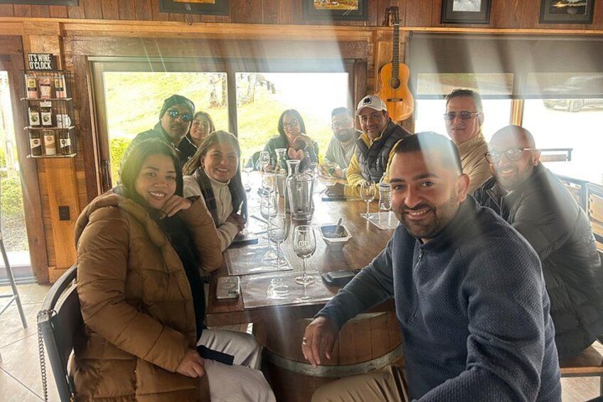 A group of people seated around a wooden table with wine glasses and bottles, in a rustic indoor setting with wooden walls and a guitar in the background. 5