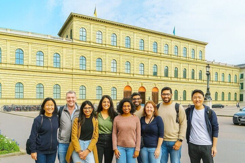 En grupp människor som står framför en stor, gul byggnad med välvda fönster och en flagga på toppen. 5