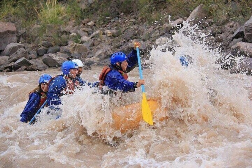Mensen in blauwe jacks en helmen peddelen met een gele kajak door een woelige rivier. 1