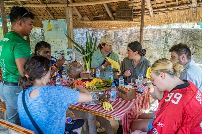Een groep mensen is samengekomen rond een tafel met eten en drinken, onder een rieten dak. 2