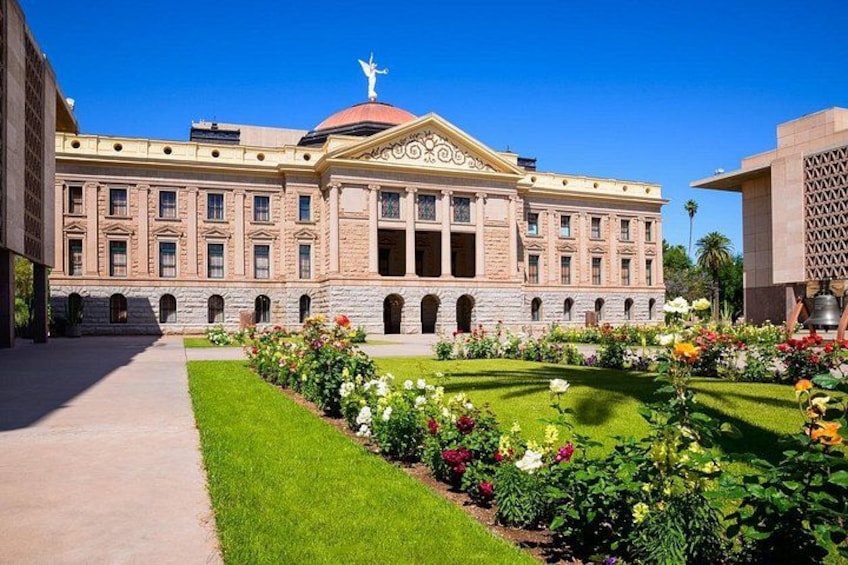 Un grand bâtiment doté d’un dôme et de colonnes, entouré d’un jardin bien entretenu où poussent différentes espèces de fleurs, le tout sous un ciel bleu azur. 2