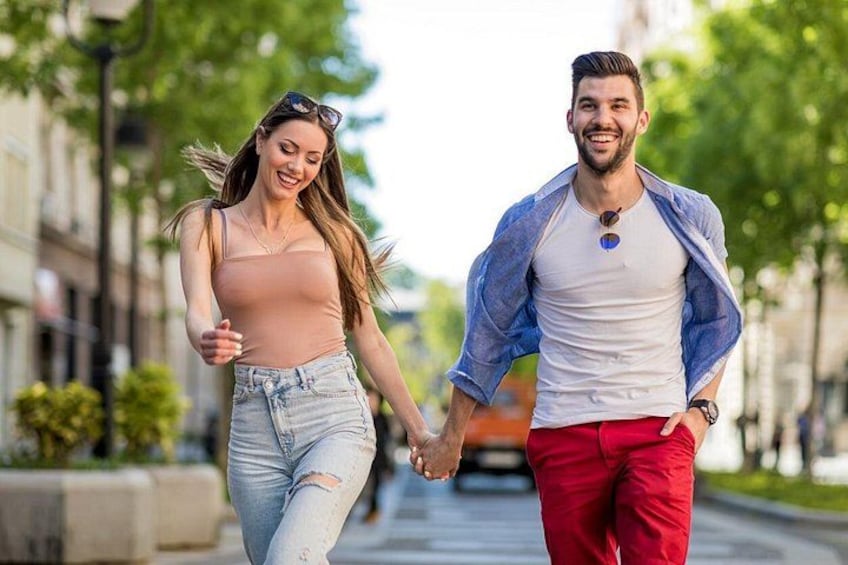 Deux personnes se promènent dans la rue, se tenant la main, avec des bâtiments et des arbres en arrière-plan. 1
