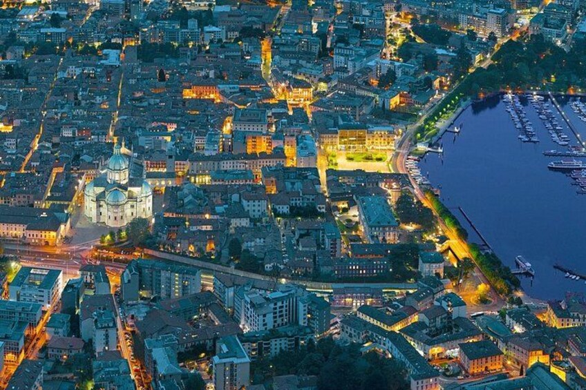 An aerial view of a city at dusk with illuminated buildings, a prominent cathedral, and a marina with boats. 2