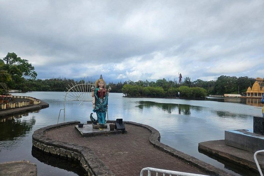 Una estatua sobre un pedestal junto a un cuerpo de agua tranquilo, rodeada de follaje y un cielo nublado. 2