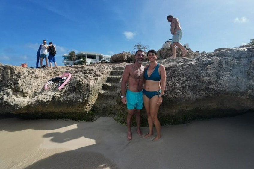 A beach with people standing on a rock formation, a person walking on the sand, and a pink object on the rock. 2