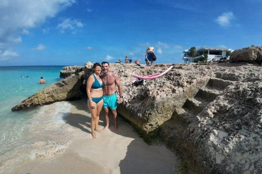 A couple standing on a rocky beach with clear turquoise water, a pink rope, and a few people in the background. 3