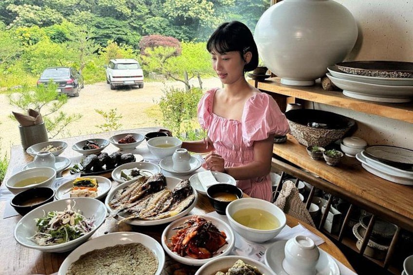 A woman preparing food at a table with various dishes and bowls, with a scenic view of trees and parked cars in the background. 2