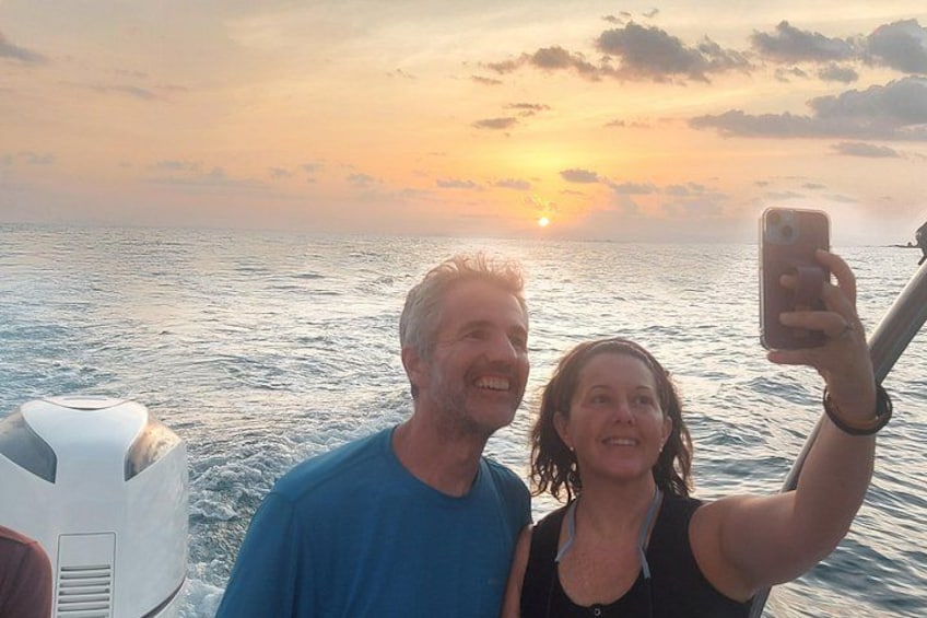 A sunset over the ocean with a boat and two people taking a selfie. 1