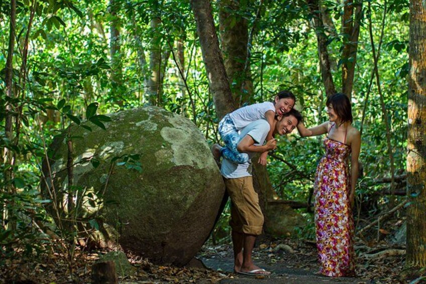 A man carrying a child on his back, a woman standing beside them, and a large rock in a forest. 5