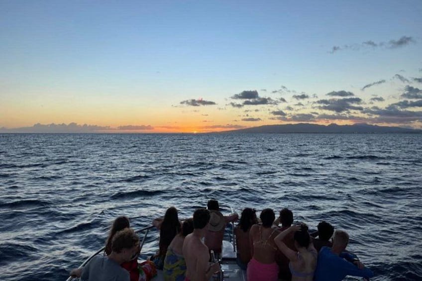 Een groep mensen op een boot bij zonsondergang, met de oceaan en een verre kustlijn op de achtergrond. 2