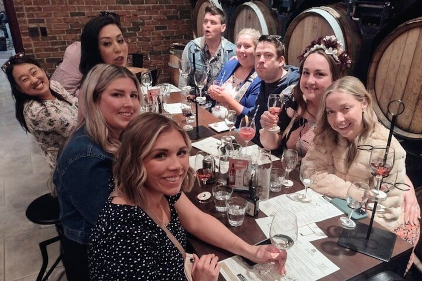 A group of people seated at a table with wine glasses, surrounded by wooden barrels. 2