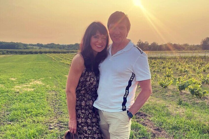 Two people standing in a field with a vineyard in the background. 3