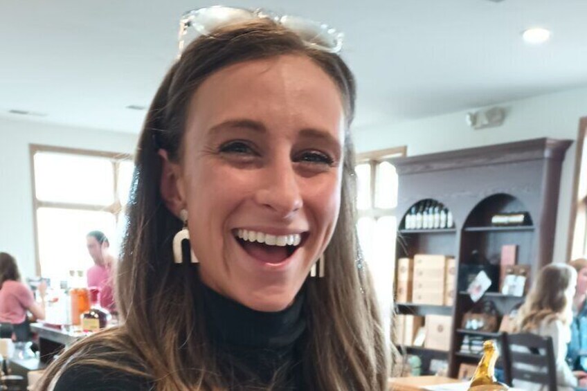 A person with long hair, wearing a black top and large earrings, sitting at a table in a restaurant with a wine rack in the background. 2