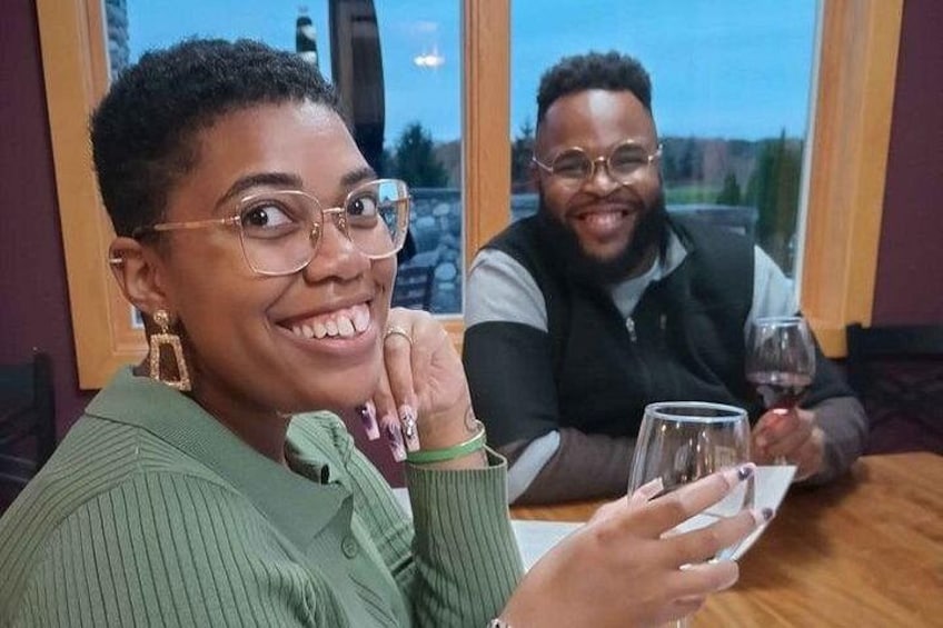 Deux personnes sont assises à une table, devant des verres à vin, dans une pièce dont la fenêtre donne sur un paysage arboré. 4