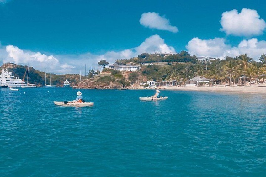A coastal scene with two kayaks on calm blue water, a beach with palm trees, and a hill with buildings in the background. 5