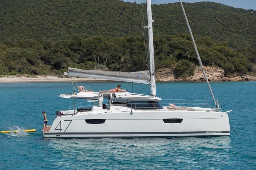 Un velero blanco con cabina y una persona en la cubierta, anclado cerca de una playa con árboles y colinas al fondo. 1