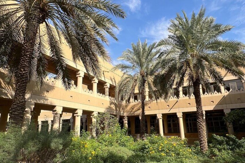 A courtyard with palm trees and a beige building with columns. 1