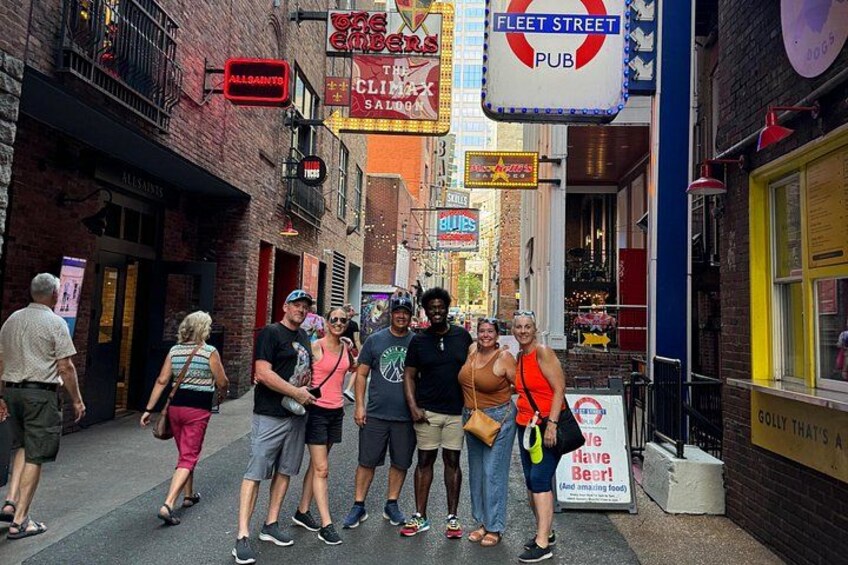 A group of people standing on a sidewalk in a narrow street with various storefronts and signs. 1
