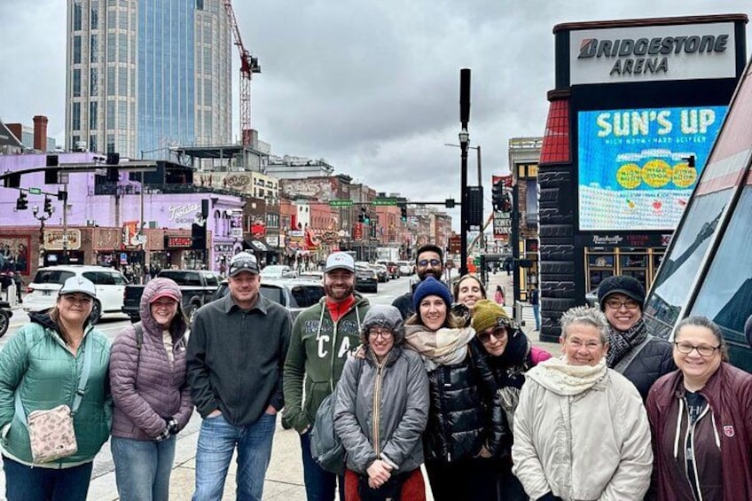 A group of people standing on a city street with a Bridgestone Arena sign and a billboard advertising 'Sun's Up.' 3