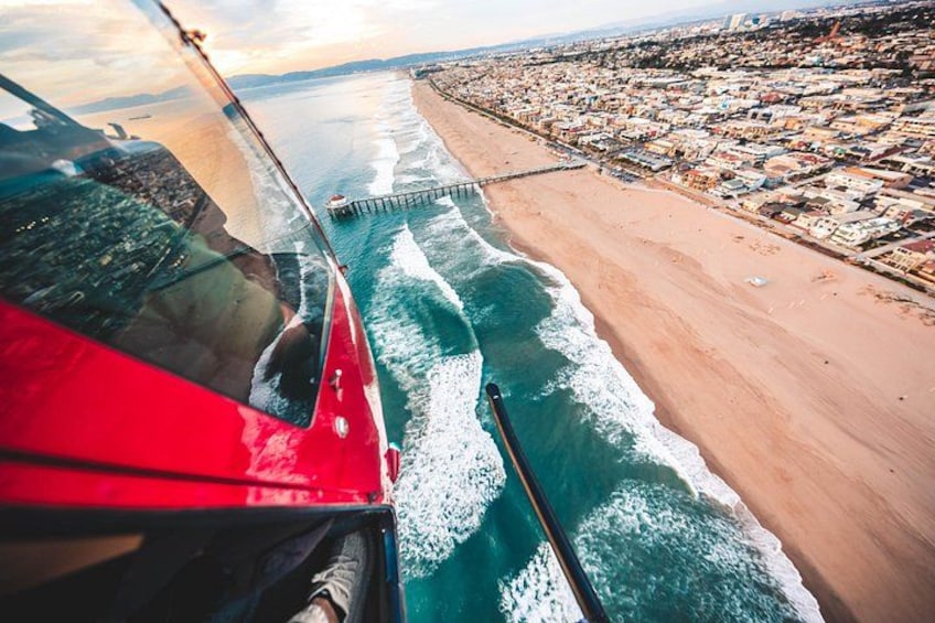 A red boat on a beach with waves, a pier, and a cityscape in the background. 1